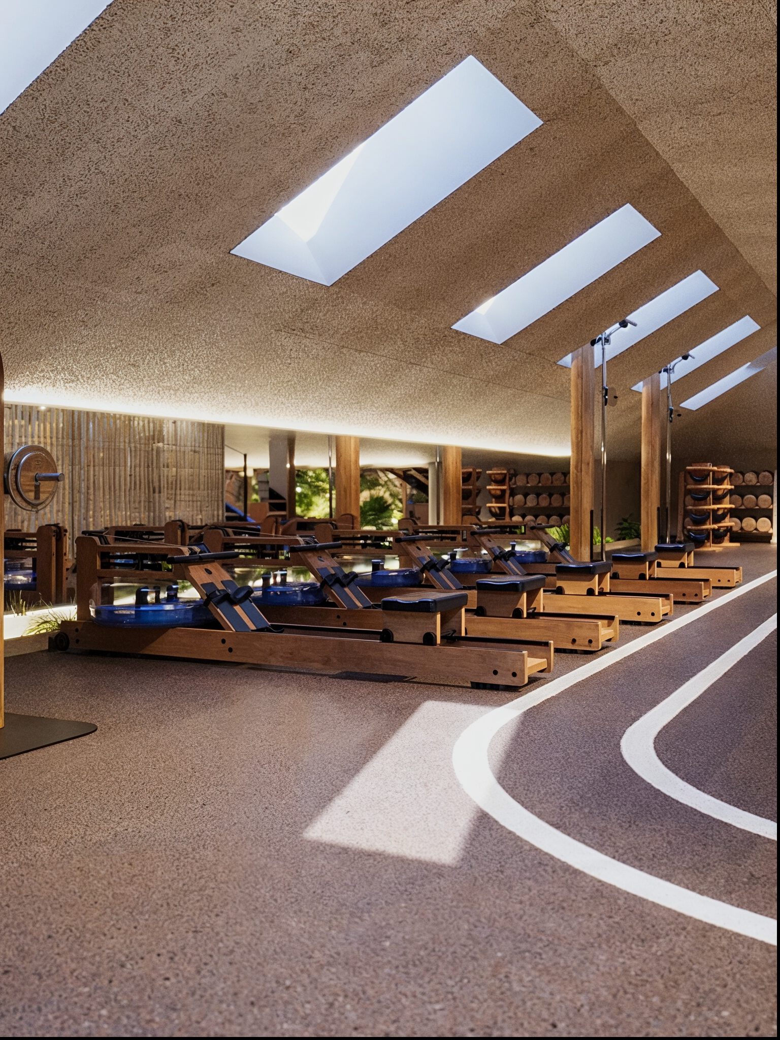Salle de sport avec plusieurs rameurs en bois alignés sous des ouvertures rectangulaires de plafond laissant passer la