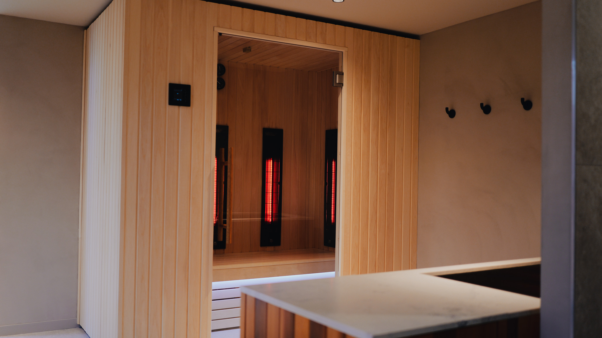 P1190005 sauna en bois clair avec porte vitrée et panneaux chauffants rouges intérieur trois patères murales noires sur mur beige